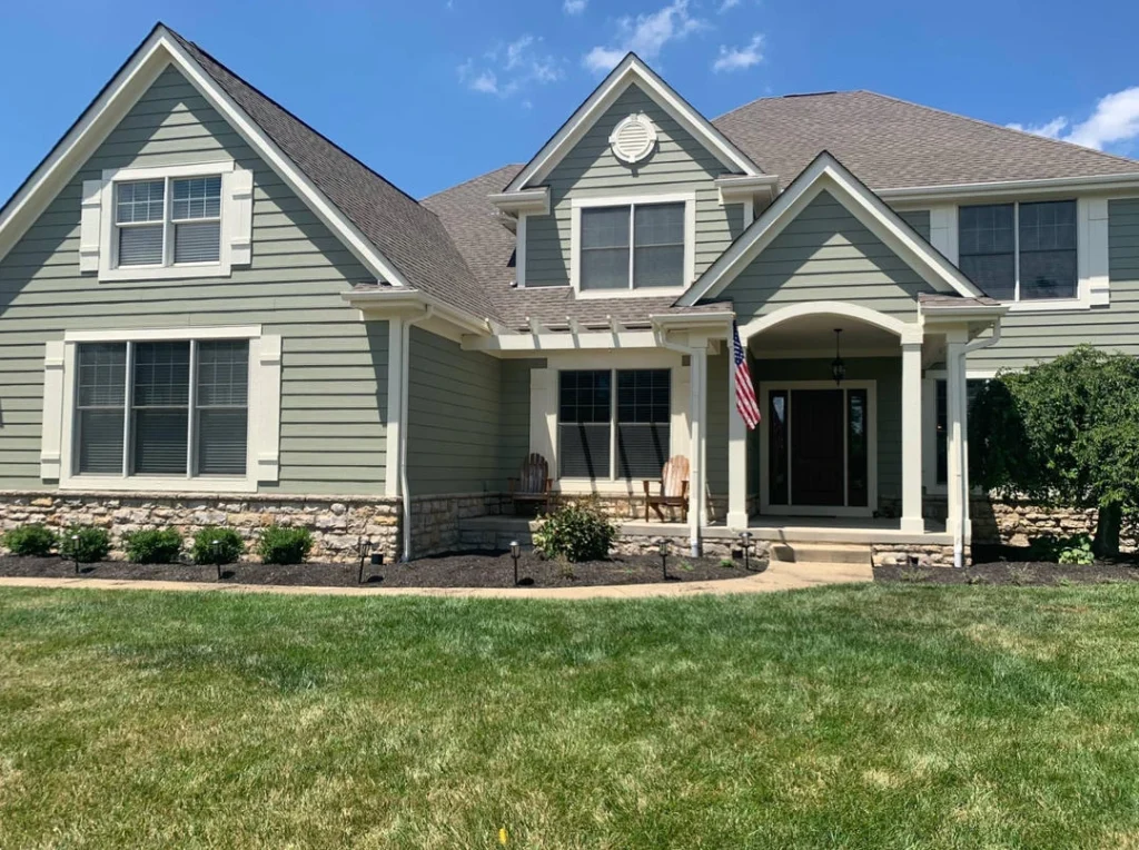 A two-story light green house with white trim, a stone foundation, and a dark brown roof. A U.S. flag hangs near the entrance, and two wooden Adirondack chairs sit on the porch.